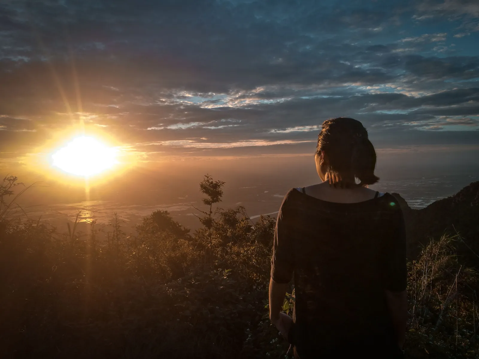 Femme de dos face à un coucher de soleil, hésitant à avancer vers l'horizon